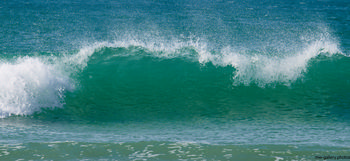 This landscape photograph captures waves breaking along the coast of Sennen Cove in Cornwall, United Kingdom. Taken during the afternoon in early spring, the image showcases the natural beauty of the beach with vibrant turquoise water and foamy crests. The waves are the central feature, demonstrating the dynamic nature of the Atlantic Ocean meeting the Cornish shoreline. The scene highlights the unspoiled coastal environment typical of southwest England, emphasizing the connection between nature and the seaside locations found in this region.